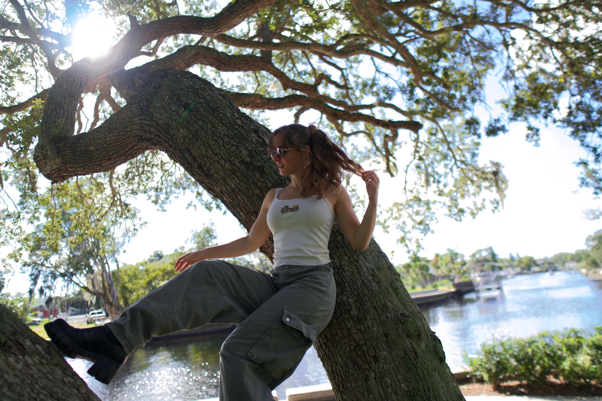 A woman seated on an oak trunk reaching out with one arm, sun flaring through the leaves