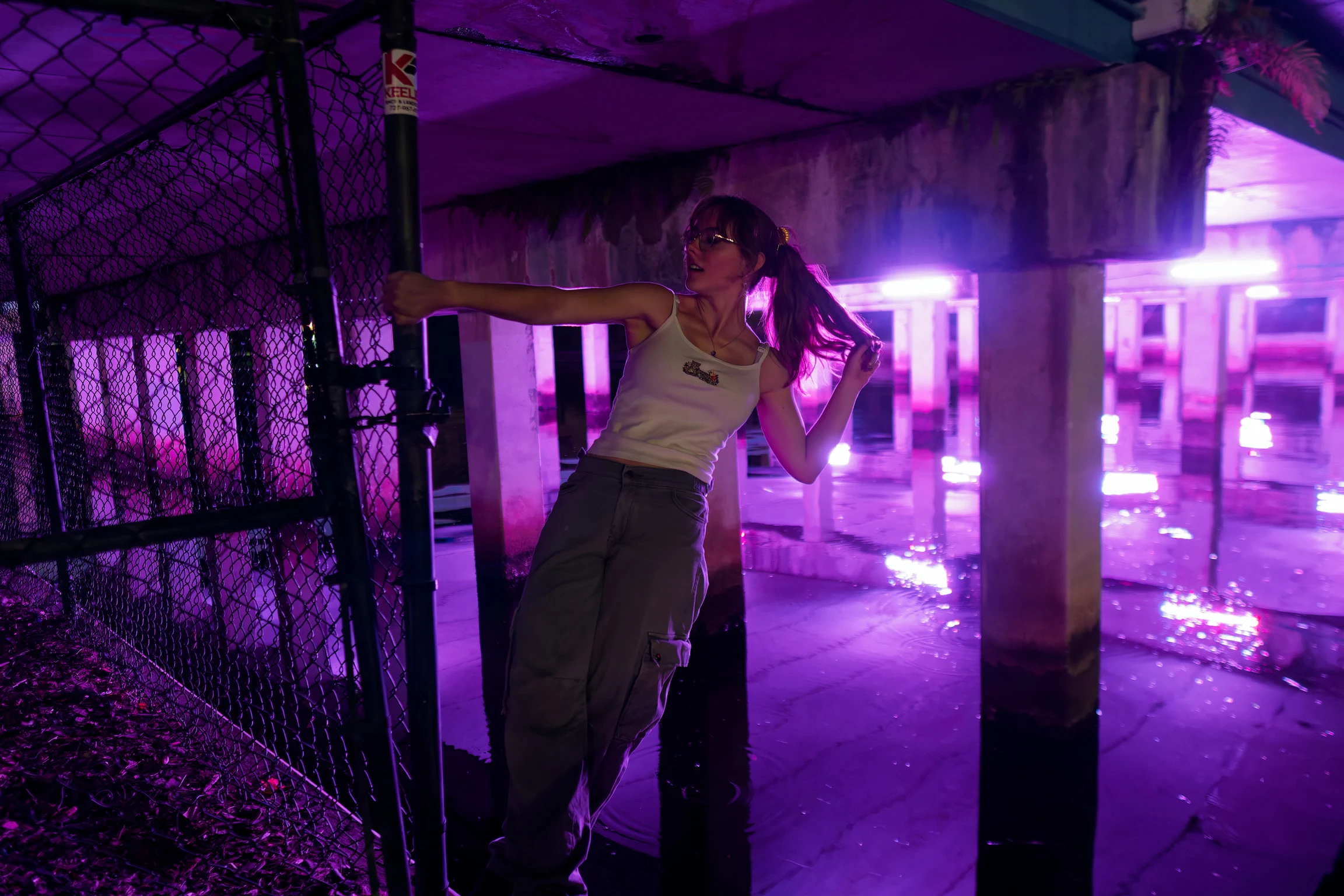 A woman leaning on a chain-link fence at night beneath a pier lit purple