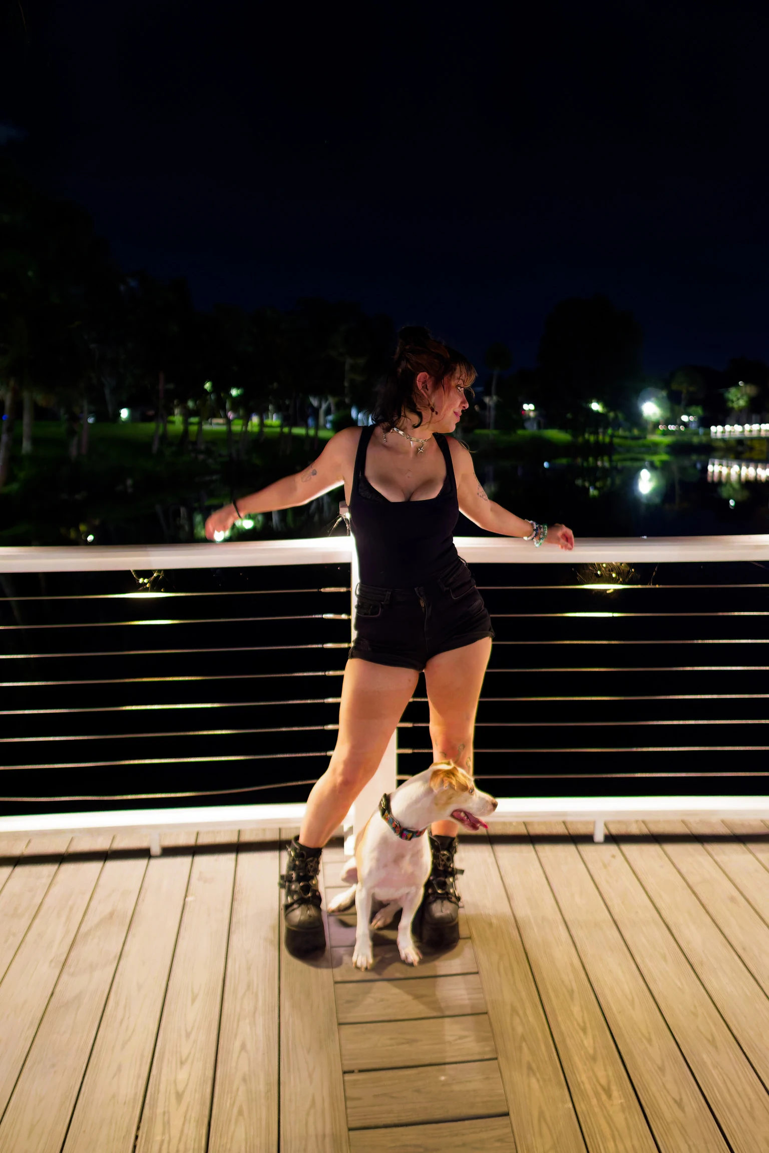 A woman in a black dress at a boardwalk railing at night with a small white dog at her feet